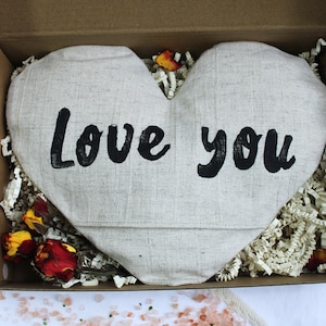 May include: A heart-shaped linen pillow with the words "Love you" printed in black. The pillow is in a brown cardboard box with white paper shreds.