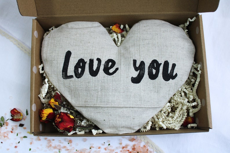 May include: A heart-shaped linen pillow with the words "Love you" printed in black. The pillow is in a brown cardboard box with white paper shreds.