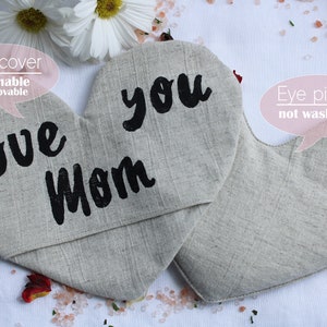 May include: Two heart-shaped eye pillows, one with the text "Love Mom" and the other with the text "you". The eye pillows are made of a light beige linen fabric. The eye pillow on the left has a removable eye cover.