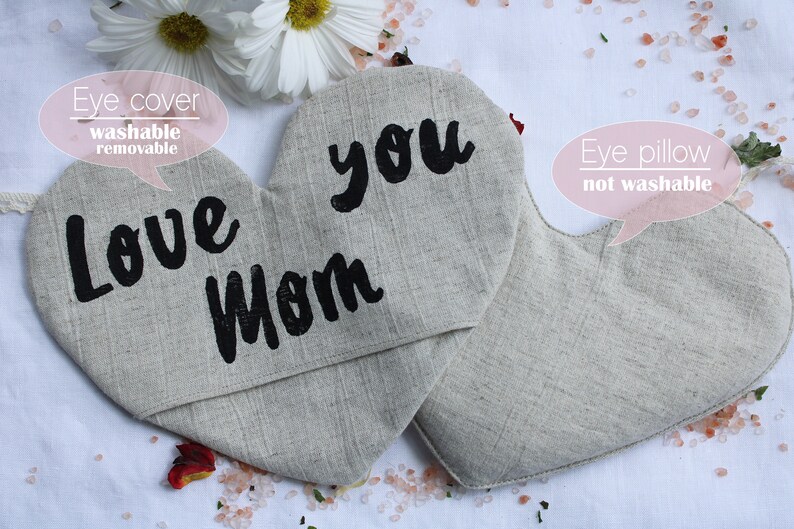 May include: Two heart-shaped eye pillows, one with the text "Love Mom" and the other with the text "you". The eye pillows are made of a light beige linen fabric. The eye pillow on the left has a removable eye cover.