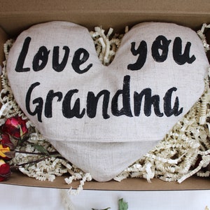May include: A heart-shaped linen pillow with the words "Love you Grandma" printed in black. The pillow is in a brown cardboard box with white tissue paper.