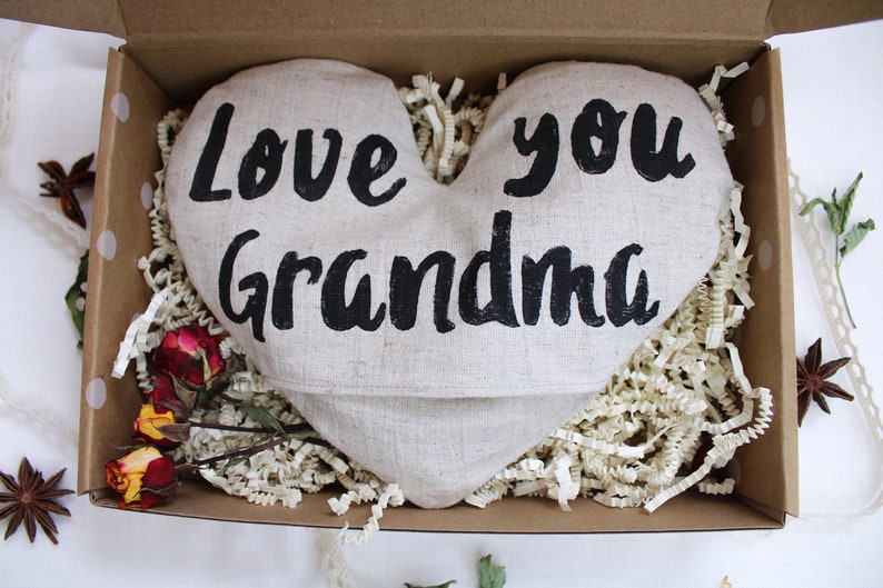 May include: A heart-shaped linen pillow with the words "Love you Grandma" printed in black. The pillow is in a brown cardboard box with white tissue paper.