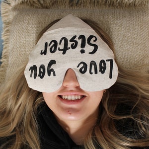 May include: A white eye mask with the text "Love you Sister!" written in black. The eye mask is on a person's face, and the person is smiling.