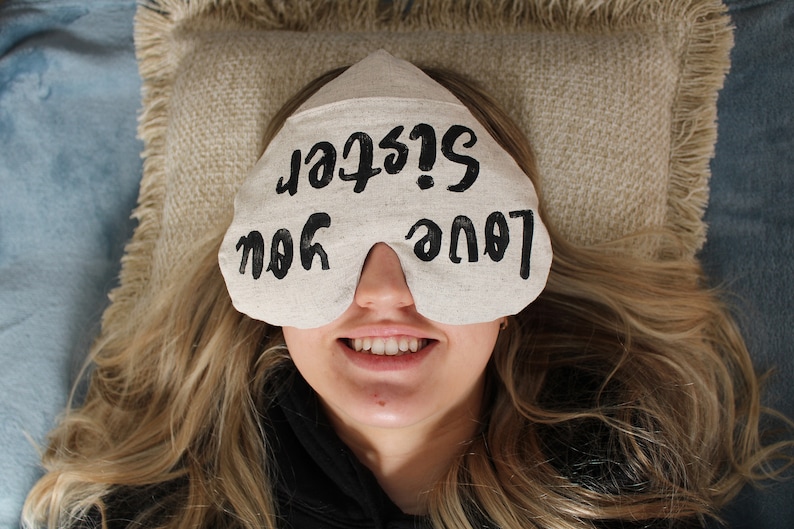 May include: A white eye mask with the text "Love you Sister!" written in black. The eye mask is on a person's face, and the person is smiling.