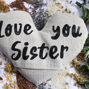 May include: A heart-shaped linen pillow with the words "Love you Sister" printed in black. The pillow is surrounded by dried herbs and flowers.