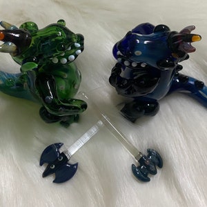 May include: Two glass dragon figurines, one green and one blue, with a clear glass axe in between them. The dragons have black eyes and white teeth.