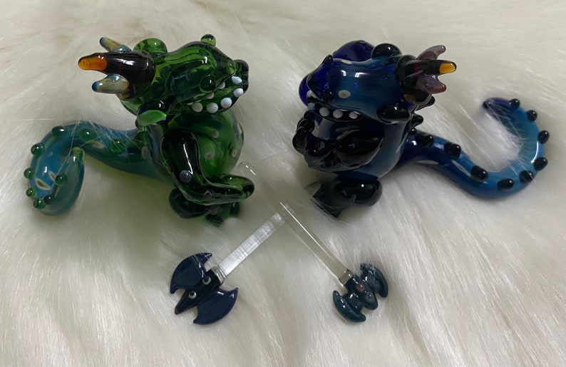 May include: Two glass dragon figurines, one green and one blue, with a clear glass axe in between them. The dragons have black eyes and white teeth.