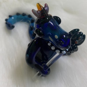 May include: A blue glass dragon figurine with a crown, white dots, and a black tail. The dragon is holding a clear glass rod in its front paws.