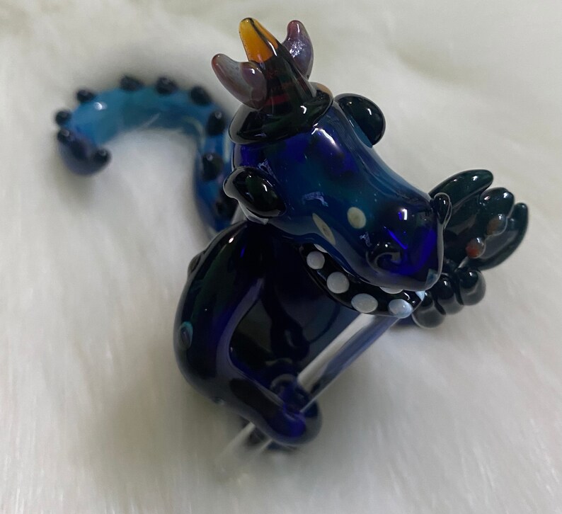 May include: A blue glass dragon figurine with a crown, white dots, and a black tail. The dragon is holding a clear glass rod in its front paws.