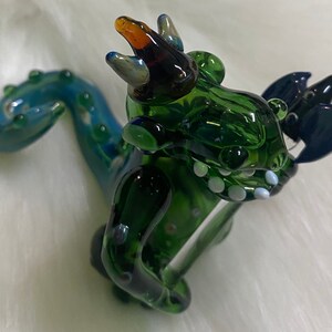 May include: A green glass dragon figurine with a blue tail and a crown. The dragon has white teeth and a blue and black wing.
