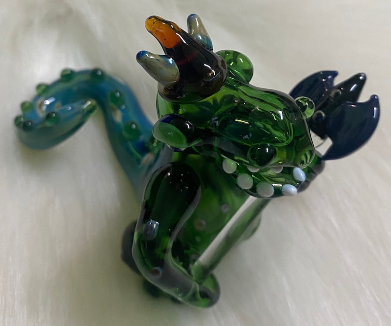 May include: A green glass dragon figurine with a blue tail and a crown. The dragon has white teeth and a blue and black wing.