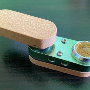 May include: A small, portable smoking pipe with a wooden lid and a metal body. The pipe has a screen and a small hole for airflow. The pipe is green and brown.