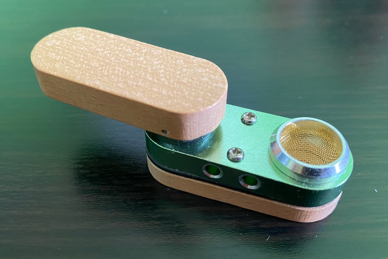 May include: A small, portable smoking pipe with a wooden lid and a metal body. The pipe has a screen and a small hole for airflow. The pipe is green and brown.