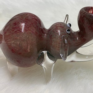 Op de afbeelding: Een glazen pijp in de vorm van een rood en wit nijlpaard met heldere glazen poten en een heldere glazen slagtand. De pijp heeft een ronde kom en een lang, dun mondstuk.