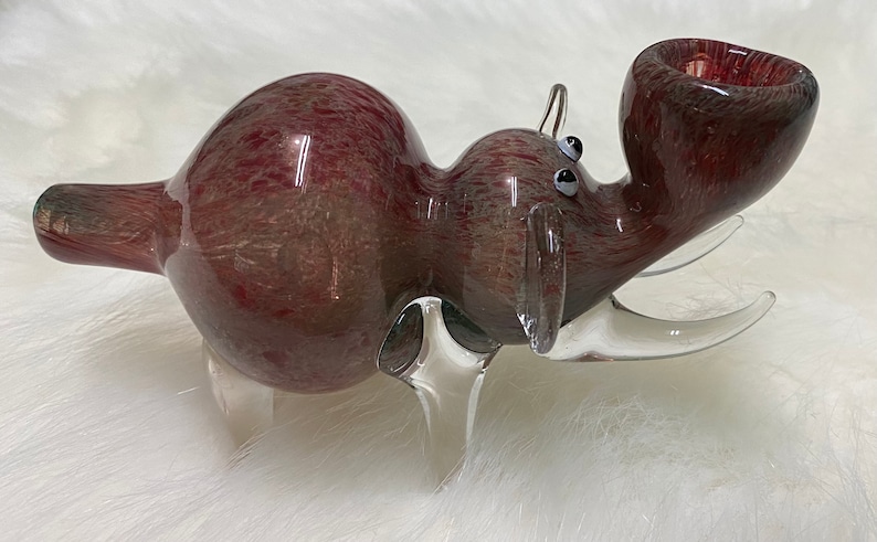 Op de afbeelding: Een glazen pijp in de vorm van een rood en wit nijlpaard met heldere glazen poten en een heldere glazen slagtand. De pijp heeft een ronde kom en een lang, dun mondstuk.