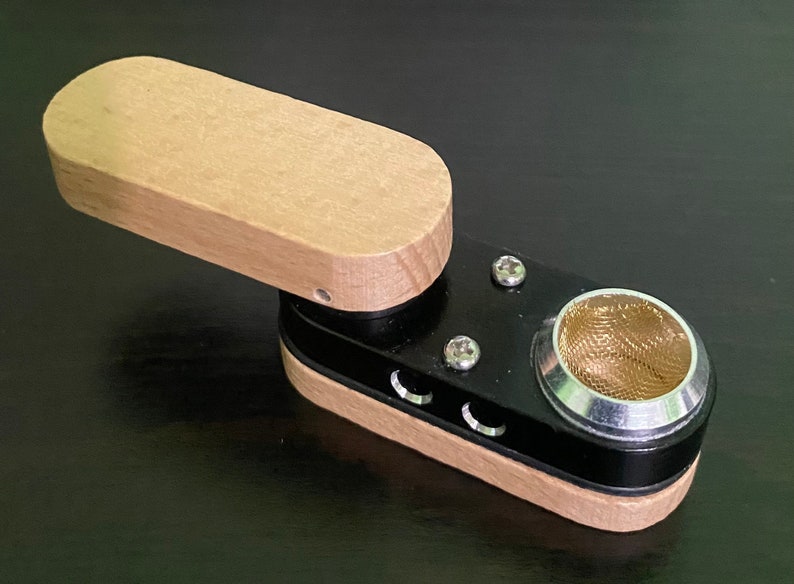 May include: A small wooden and black pipe with a metal bowl and a screen. The pipe has a hinged lid and three small holes on the side.