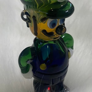 May include: A green and blue glass figurine of a character wearing a green hat with a white letter L on it. The figurine has a yellow face with black eyes, a black moustache, and a black mouth.