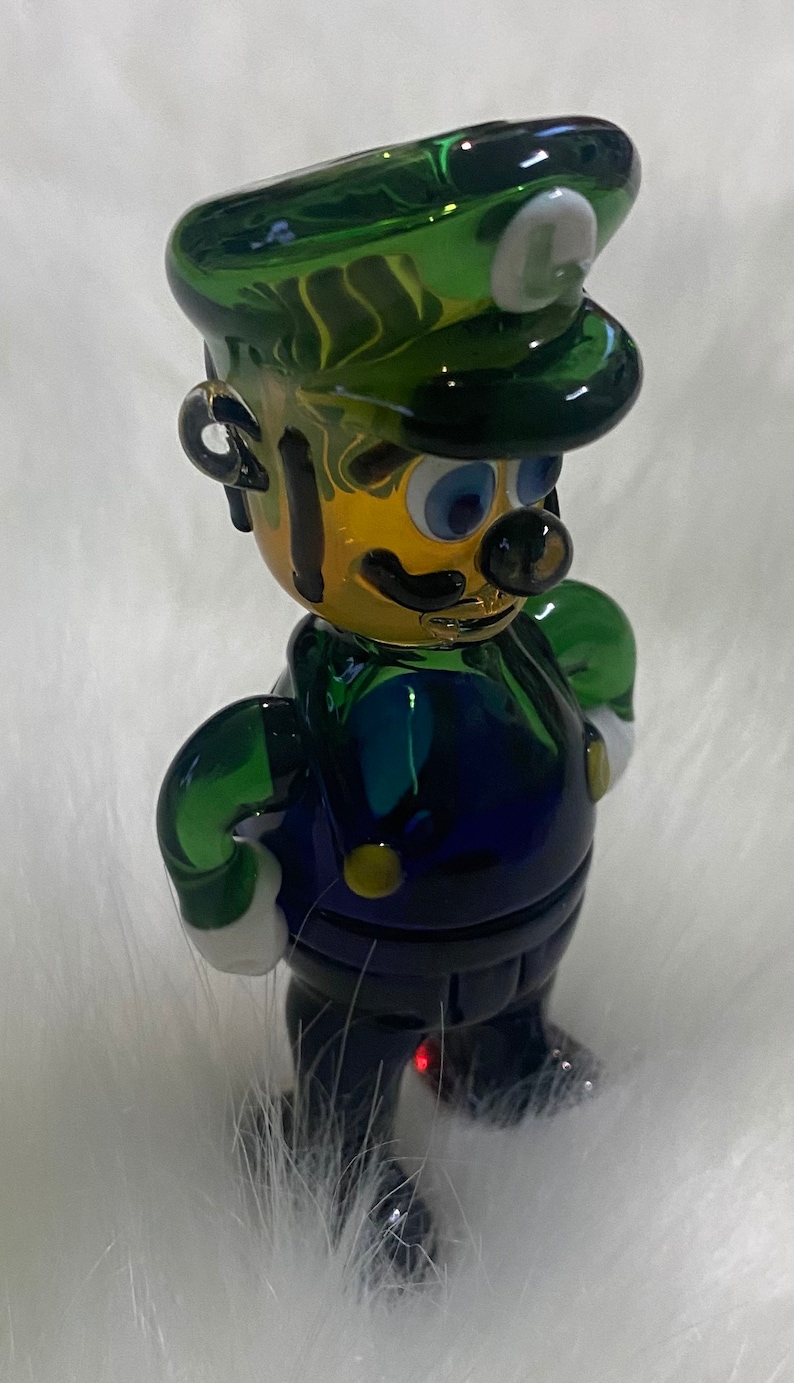 May include: A green and blue glass figurine of a character wearing a green hat with a white letter L on it. The figurine has a yellow face with black eyes, a black moustache, and a black mouth.