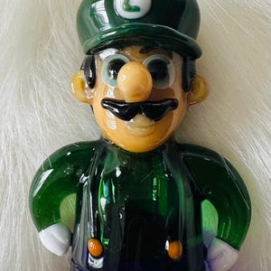 May include: A green glass figurine of a video game character wearing a green hat with a white "L" on it. The character has a brown moustache, a brown nose, and large blue eyes. The figurine is wearing a green shirt and blue trousers.