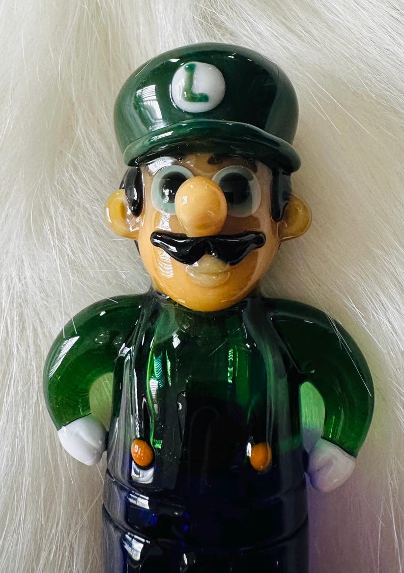 May include: A green glass figurine of a video game character wearing a green hat with a white "L" on it. The character has a brown moustache, a brown nose, and large blue eyes. The figurine is wearing a green shirt and blue trousers.