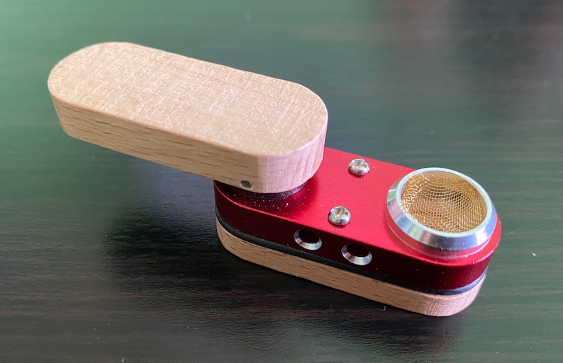 May include: A small, wooden and red metal pipe with a screen on the end. The pipe has a hinged lid and a small hole on the side.