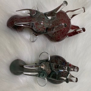 Op de afbeelding: Twee glazen dierenfiguren, een rood en een grijs, met een gespikkelde afwerking. Beide hebben lange, dunne poten en een rond lichaam.