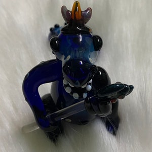May include: A blue glass dragon figurine with a black and white spotted design. The dragon is wearing a crown and holding a sword.
