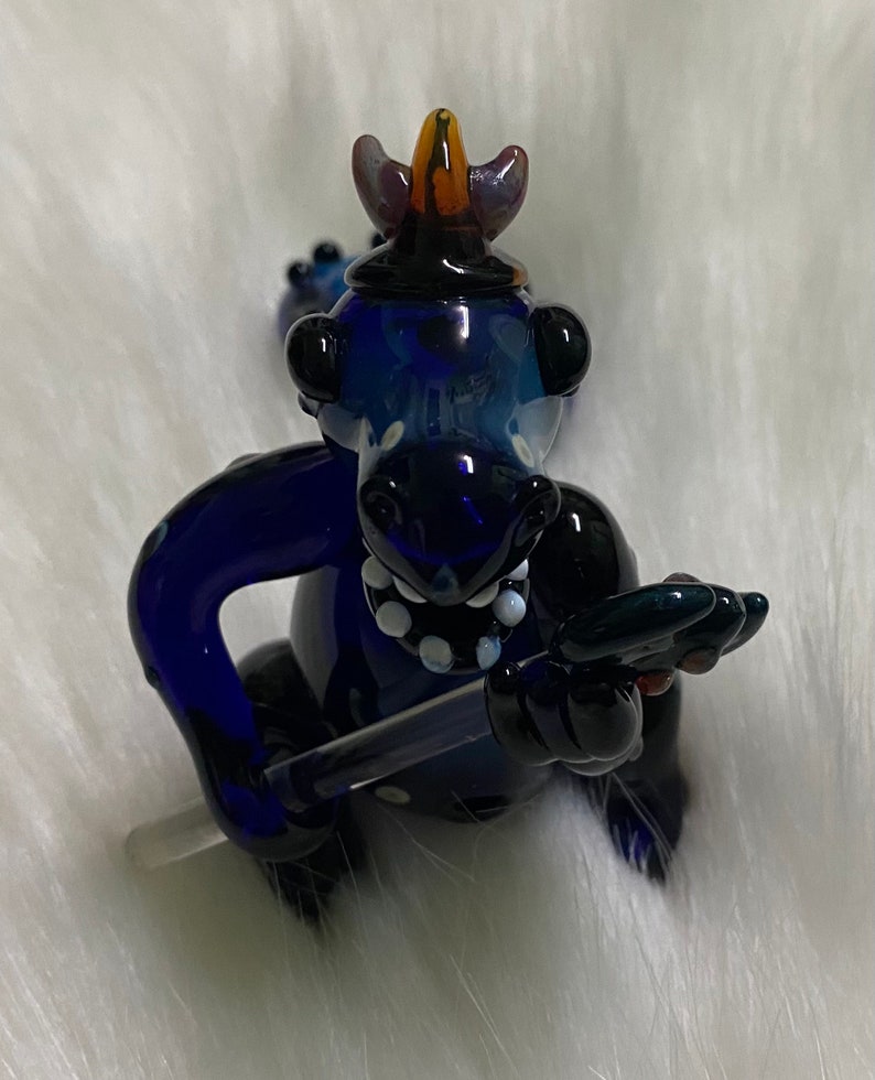 May include: A blue glass dragon figurine with a black and white spotted design. The dragon is wearing a crown and holding a sword.