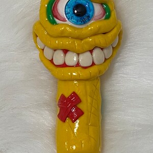 May include: A yellow ceramic pipe shaped like a monster with a single blue eye, red and green eyelids, and a wide toothy grin. The pipe has a red X on the side.