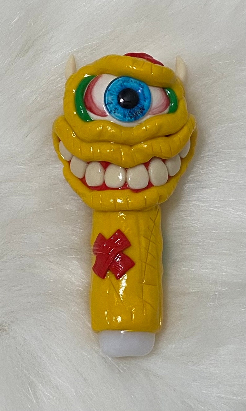 May include: A yellow ceramic pipe shaped like a monster with a single blue eye, red and green eyelids, and a wide toothy grin. The pipe has a red X on the side.