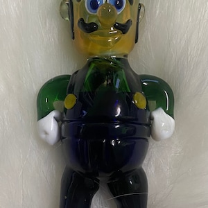 May include: A glass figurine of a character wearing a green hat with a white "L" on it. The character is made of green, blue and yellow glass and has a black moustache and blue eyes.