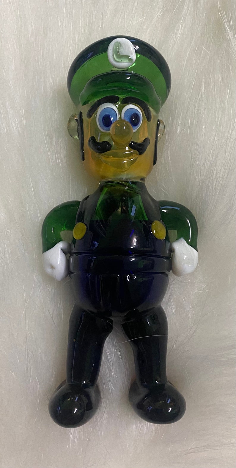 May include: A glass figurine of a character wearing a green hat with a white "L" on it. The character is made of green, blue and yellow glass and has a black moustache and blue eyes.