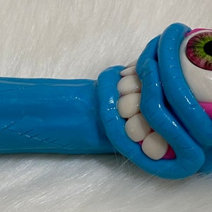 May include: A blue and pink ceramic pipe shaped like a monster with a single large eye and a toothy grin.