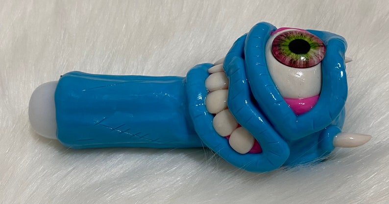 May include: A blue and pink ceramic pipe shaped like a monster with a single large eye and a toothy grin.
