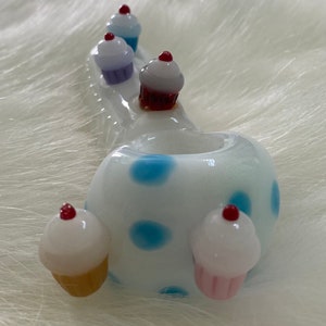 May include: White glass pipe with blue polka dots and cupcake accents. The pipe has a bowl and a stem. The cupcakes are decorated with red, white, and pink icing.