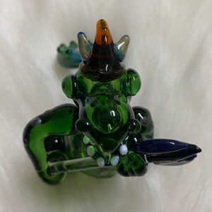 May include: Green glass dragon figurine with a blue and orange hat, holding a blue glass sword. The dragon has white dots on its body.