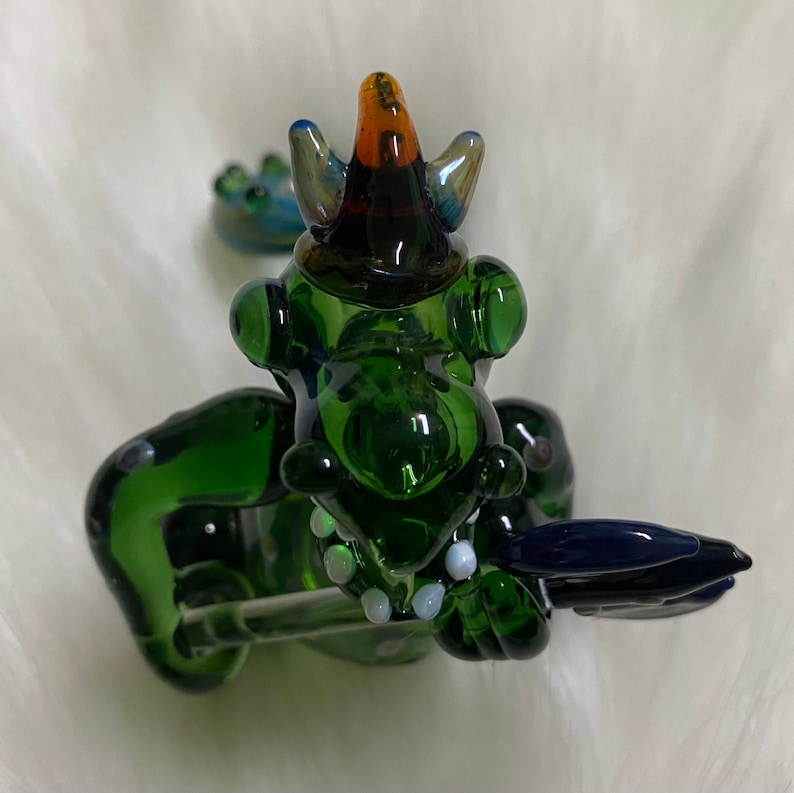 May include: Green glass dragon figurine with a blue and orange hat, holding a blue glass sword. The dragon has white dots on its body.
