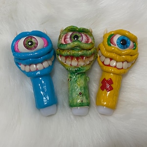 May include: Three ceramic figurines with cartoon monster faces. Each figurine has a single large eye and a toothy grin. The figurines are blue, green, and yellow.