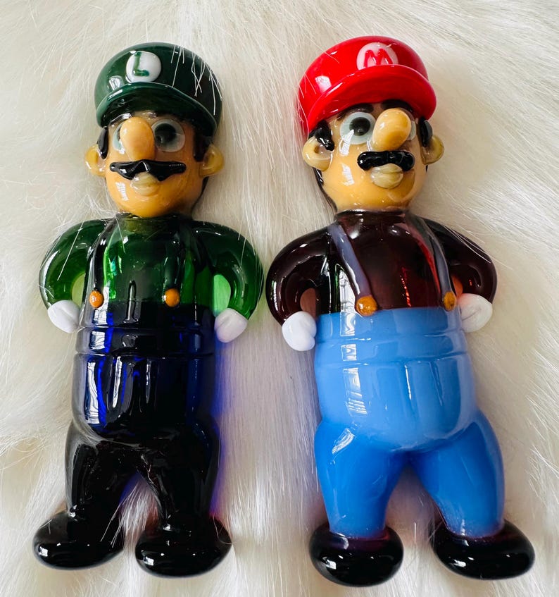 May include: Two glass figurines of Mario and Luigi from the Super Mario Bros. video game series. The figurines are hand-painted and feature detailed facial expressions and clothing. Mario is wearing his signature red hat and blue overalls, while Luigi is wearing a green hat and blue overalls.