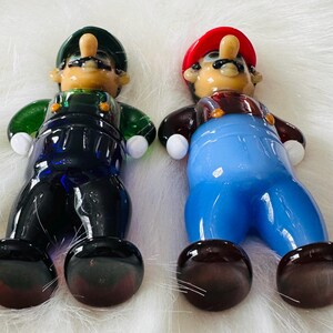 May include: Two glass figurines of Mario and Luigi from the Super Mario Bros. video game series. Mario is wearing a red hat and blue overalls, while Luigi is wearing a green hat and blue overalls. Both figurines have brown shoes and white gloves.