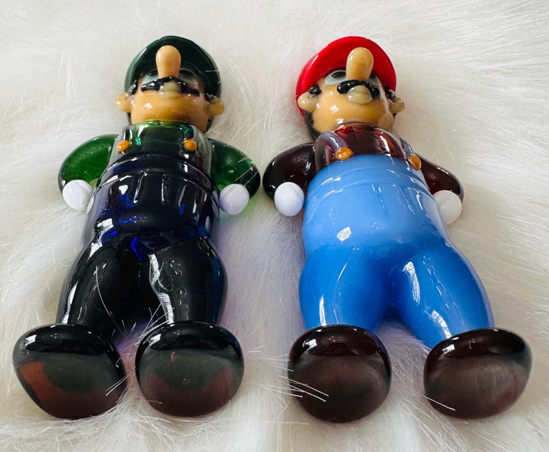 May include: Two glass figurines of Mario and Luigi from the Super Mario Bros. video game series. Mario is wearing a red hat and blue overalls, while Luigi is wearing a green hat and blue overalls. Both figurines have brown shoes and white gloves.