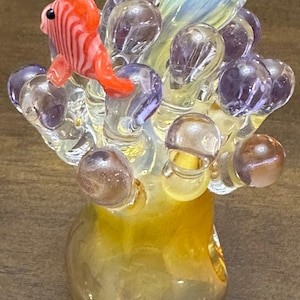 Peut inclure: Sculpture en verre artisanal représentant un poisson rayé orange et blanc sur un amas de formes claires en forme de bulles. La base est jaune doré, avec des accents violets et transparents.