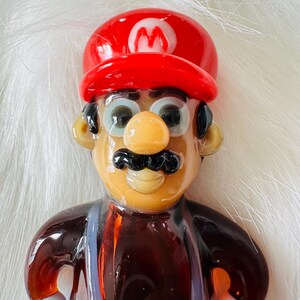 May include: A glass figurine of a cartoon character wearing a red hat with a white 'M' on it. The character has a brown moustache, brown eyes, and a brown nose. The figurine is made of clear glass with brown, red, and white accents.