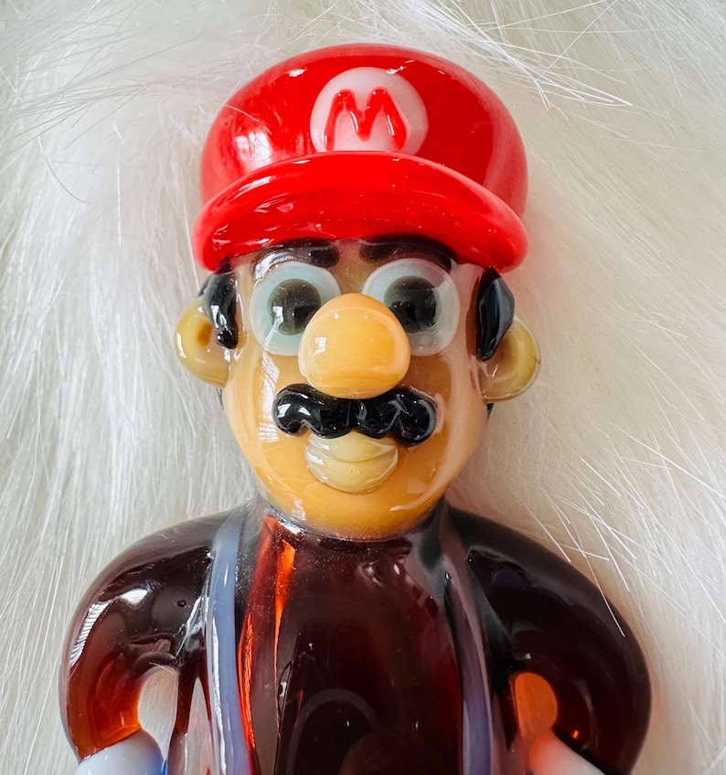May include: A glass figurine of a cartoon character wearing a red hat with a white 'M' on it. The character has a brown moustache, brown eyes, and a brown nose. The figurine is made of clear glass with brown, red, and white accents.