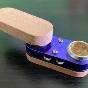 May include: A small, portable wooden and metal smoking pipe with a blue metal body and a wooden lid. The pipe has a mesh screen and a small hole for airflow.