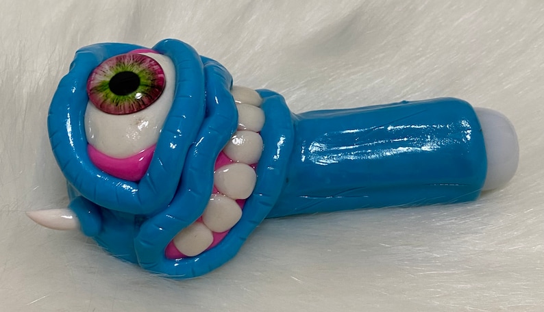 May include: A blue and pink ceramic pipe with a single large eye and a row of teeth. The pipe has a white tip.