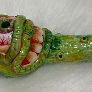 May include: A green and yellow ceramic pipe shaped like a monster with a large eye and teeth. The eye is red and has a black pupil.