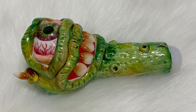 May include: A green and yellow ceramic pipe shaped like a monster with a large eye and teeth. The eye is red and has a black pupil.