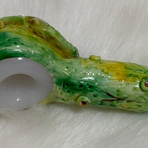 May include: A green and yellow ceramic pipe shaped like a tree log with a white bowl. The pipe has a small hole on the side and a brown stem.