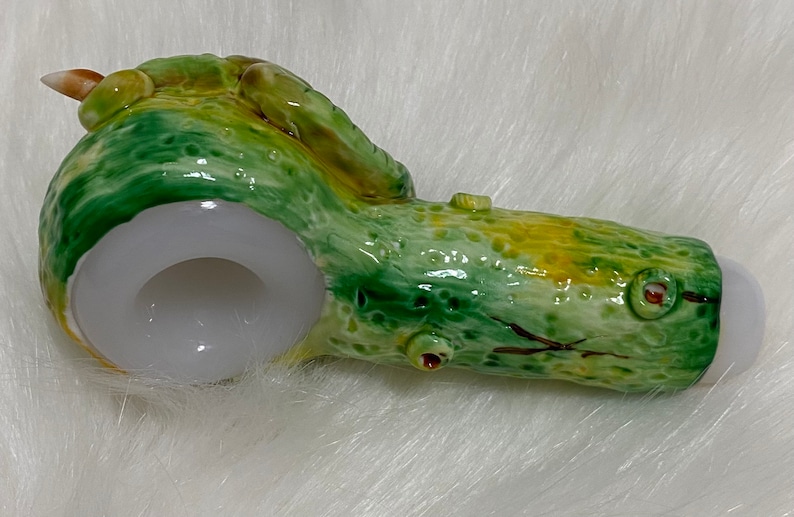 May include: A green and yellow ceramic pipe shaped like a tree log with a white bowl. The pipe has a small hole on the side and a brown stem.